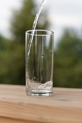 A stream of water flowing into an empty transparent glass standing on a wooden board.