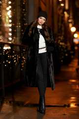 Fashionable stylish woman in black fur coat walking at city street at night. Christmas decoration at background