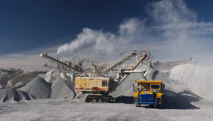 Heavy excavator and dumper during crushed stone loading on the background of a rock stone crushing plant and a blue sky on a sunny day, panorama. Mining industry. Heavy equipment.