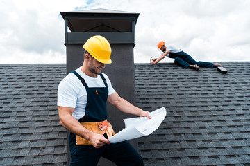 selective focus of handsome builder in helmet holding blueprint near colleague repairing roof