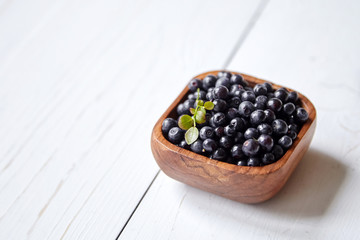 Bilberries with green leaf into a wooden bowl on white table. Many fresh dark blue berries on wooden background with copy space