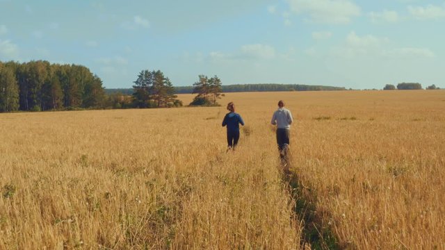 A young man and a girl with sports figures in sportswear are running along a cereal field. Summer day. In the background a blue sky with clouds and a forest.