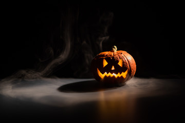 Halloween, orange pumpkin with a scary luminous face on a dark background. Thick gray smoke comes...