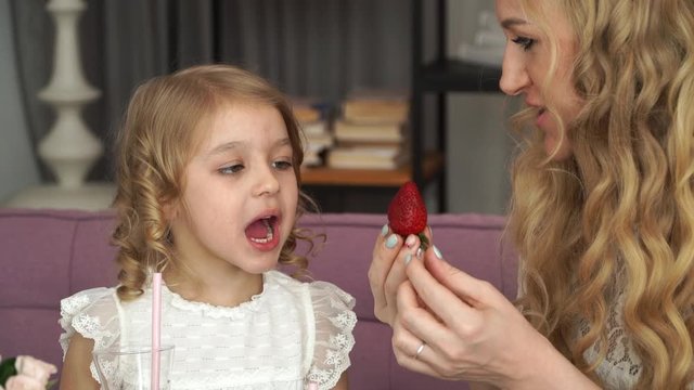 Young smiling mother and little daughter eat red ripe strawberries. Woman gives taste of girl’s strawberries and tastes sweet berry itself. Happy family