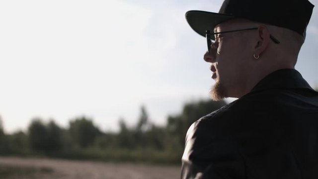 Back view on the caucasian male dancer in sunglasses walking on the road. Tough guy in a black leather jacket. Following shot of the traveling man. Active lifestyle. Sunny day 4K Close up
