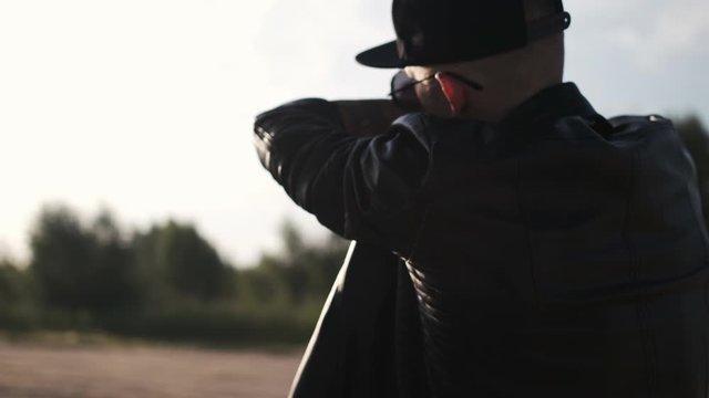 Portrait shot of young man walking on the road. View from the back. Caucasian male dancer in sunglasses. Tough guy in black leather jacket. Following shot of the traveling man. Krump. Hip hop style