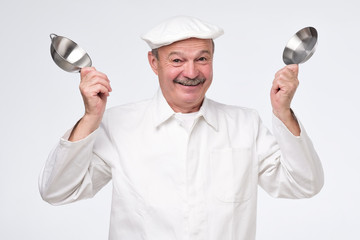 Mature hispanic man in white unifrom with fummy small kitchen pans. Studio shot