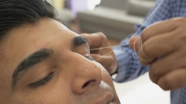 Skillful beautician makes threading hair removal procedure. Ancient Asian method of eyebrow hair removal. Threading, epilation technique in hair salon. Handsome customer makes depilation of eyebrows.