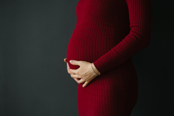 A pregnant woman in a Burgundy dress holds her hands on her stomach on a dark gray background. The...