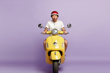 Emotional man driver poses on yellow motorbike, wears protective headgear and white t shirt, shocked with very high speed on his route, isolated over violet background. Reaching destination.