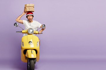 Pizza delivery courier carries stack of carton boxes over head, wears protective helmet and white t shirt, rides on yellow motorbike, poses against purple background, blank space. Food transportation