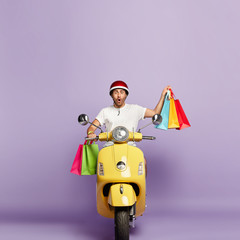 Vertical image of impressed handsome guy in casual wear, returns from shop, carries shopping bags, shocked, drives on fast motorbike, works as courier, isolated over purple background. Purchasing