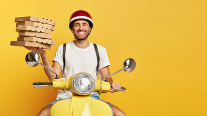Pizza delivering concept. Positive young man with glad expression, wears helmet, white t shirt, poses on scooter with carton boxes conveys fast food from restaurant. Deliveryman uses company motorbike