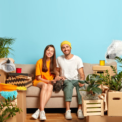 Photo of happy pleased family couple pose on sofa with french bulldog, relax on sofa together after tired moving day, enjoy having new home, many boxes with household stuff around in empty room