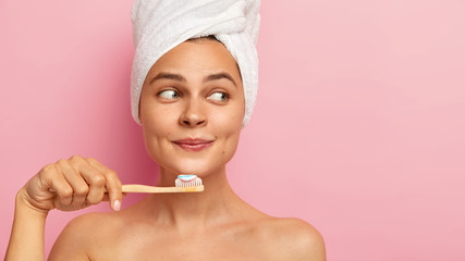 Close up shot of calm relaxed woman holds toothbrush, cleans teeth after taking bath, wears wrapped white towel on head, has naked body, healthy clean skin, focused aside, isolated on pink wall
