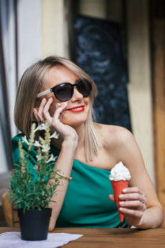 beautiful blonde girl in sunglasses, green dress and with red lipstick sits at table of street cafe. Woman holding a red ice cream cone and talking on a cell phone