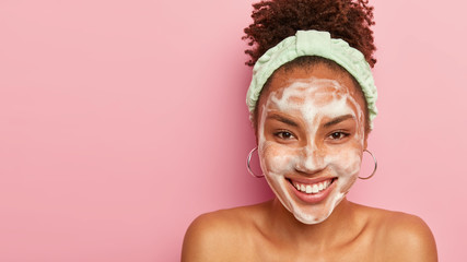 Pretty overjoyed dark skinned lady uses facial foam for washing cosmetic off on face, has broad...