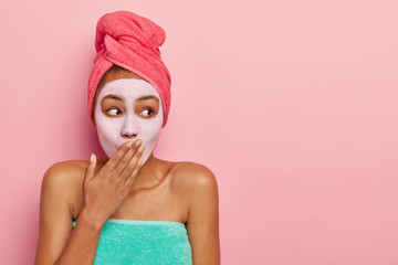 Horizontal shot of surprised African American woman covers mouth, applies nourishing mask for removing dead cells, wears wrapped towel on head, stands against pink background. Skin care concept