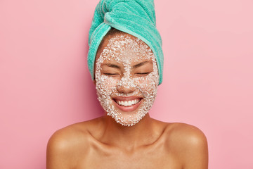 Headshot of pretty smiling woman applies salt granules on face, keeps eyes closed, shows white...