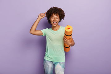Healthy lifestyle and body care concept. Happy overjoyed woman holds yoga mat, raises arm, dressed in casual t shirt and leggings, being pleased by active workout, isolated on purple background