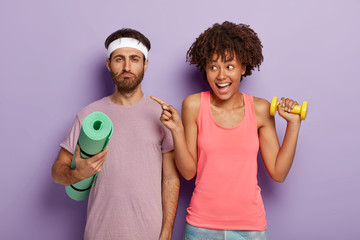 Serious unshaven man holds karemat, ready for training, cheerful African American woman points aside with index finger, holds dumbbell, works on muscles, stand together in gym. Sport concept