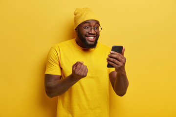 Happy joyous black man clenches fist with triumph, watches football match online, focused in...