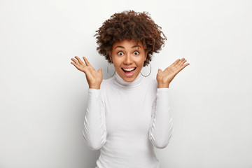 Overemotive dark skinned young woman reacts on unexpected surprise, spreads palms raised up, grins happily at camera, wears round earrings and casual turtleneck, isolated over white background