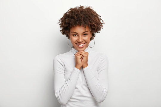 Horizontal View Of Pretty Crisp Girl With Afro Hair, Keeps Both Hands Together Under Chin, Has Glad Expression, Wears Round Earrings, Poloneck Jumper, Isolated On White Background, Enjoys Spare Time