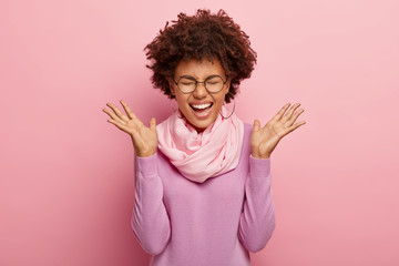 Photo of cheerful woman with curly hair, laughs with pleasure, speads palms sideways, wears silk scarf and purple poloneck, optical spectacles for vision correction, feels happy and impressed.