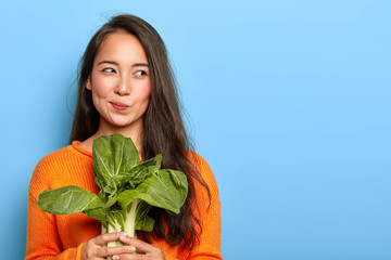 Photo of attractive young woman holds fresh green vegetable, eats healthy food at home, uses food product for making vegetarian salad, wears orange jumper, poses indoor. Home growing concept.