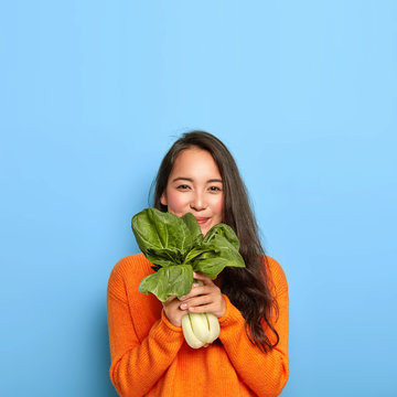 Satisfied Korean woman with pleased face expression, buys healthy dieting food, holds raw green vegetable, going to make tasty vegetarian lunch, wears casual orange jumper, poses over blue wall