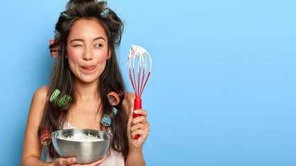 Pleased dark haired woman licks lips, enjoys sweet taste of cream, holds whisk and bowl, mixes...