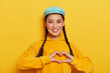 Young happy Korean woman makes heart gesture over chest, has two pigtails, wears blue cap and yellow hoody, expresses good emotions, shows love to someone, poses indoor. My heart is for you.
