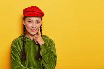 Image of lovely girl with Asian appearance, minimal makeup, touches cheek with index finger, looks positively, enjoys spending free time on shopping, wears red beret, green sweatshirt, stands indoor