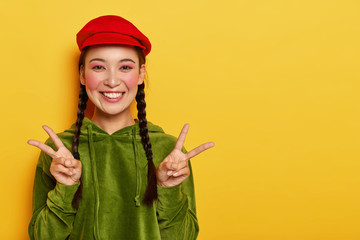 Smiling glad pretty Asian teenage girl shows peace gesture with both hands, smiles broadly, stands...