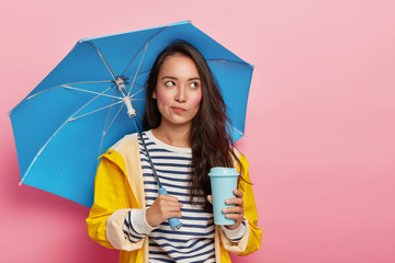 Pensive young woman with Asian appearance, walks during rainy cloudy day under umbrella, drinks...