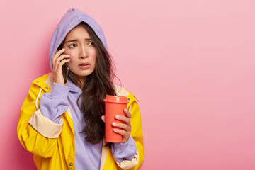 Displeased Asian woman with rosy cheeks, has nervous face expression, calls boyfriend via smartphone, tries to warm herself with takeaway coffee, wears yellow raincoat and hood, poses indoor