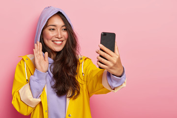 Smiling pleasant looking Asian girl waves palm and says hello to camera of modern smartphone, makes video call, has long dark hair, wears purple sweatshirt and yellow raincoat, poses indoor.