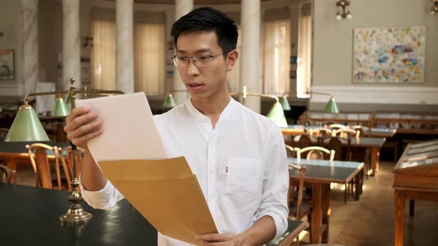 Portrait of young asian male student excitingly opening envelope with exam results in library of university