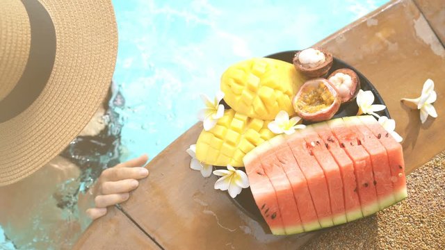 4K beautiful arrangement of multicolored tropical fruit near outdoor pool with woman swimming by and taking mangosteen