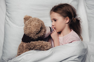 Child showing hush sign while lying in bed with teddy bear