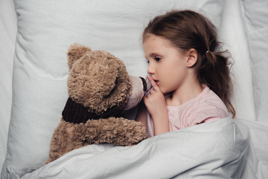 Child Showing Hush Sign While Lying In Bed With Teddy Bear