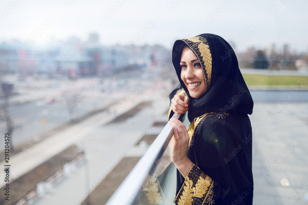 Wall mural Gorgeous muslim woman in traditional wear looking at beautiful sight while standing on rooftop.