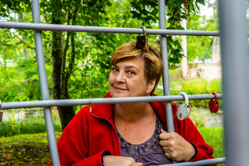 Fototapeta premium Mature woman in red jacket is resting in the Park by the river. Bridge with locks from lovers.