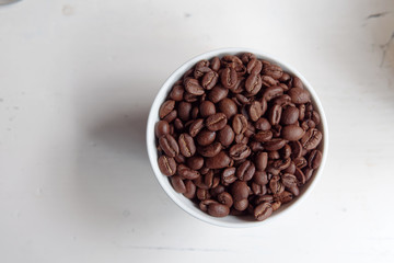 Natural coffee beans heap in white bowl on white wooden table, top view.