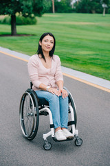 young disabled woman in wheelchair smiling at camera while walking in park