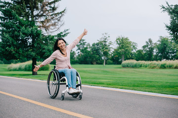 Fototapeta premium happy disabled woman gesturing and looking at camera while sitting in wheelchair in park