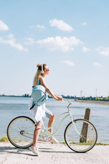 Obraz na płótnie Canvas side view of girl with backpack biking in headphones near river in summer