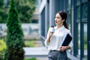 Fototapeta premium attractive journalist in formal wear holding microphone and speaking outside
