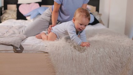 Happy mom in pajamas hugs her one-year-old baby in the bedroom on the bed in the morning. Parent and little kid relaxing at home. Mother and child on a white and grey bed.